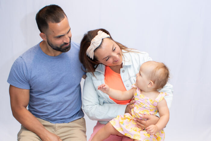 A dad and mom sit on stools with the baby daughter on mom's lap. The baby is looking up at her parents and they are looking at her.