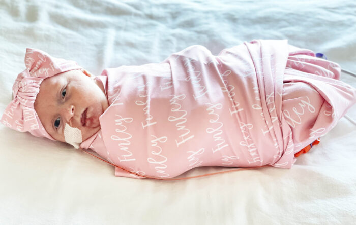 A newborn girl is wrapped in a pink blanket that says Finley. She's earing a hat with a bow and has a tube in her nose. 