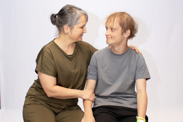 A mom and son sit next to each other on stools. They are looking at each other. The mom has one hand on his shoulder and the other on his arm.