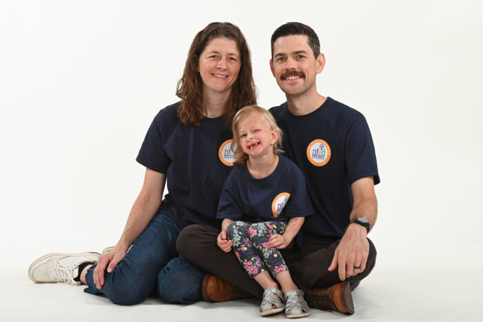 A mom and dad sit on the floor with their preschool aged daughter on the dad's lap.