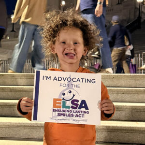 A young boy wearing an orange shirt with curly hair holds a sign saying I'm Advocating for ELSA. He's standing on the steps of the U.S. Capitol building.