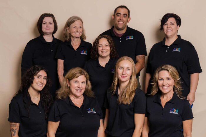 The staff are posed in two rows in front of a neutral background. The staff are all wearing black shirts.