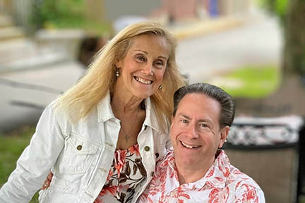 Pamela is sitting on her husband's  lap. Both are wearing Hawaiian style shirts.