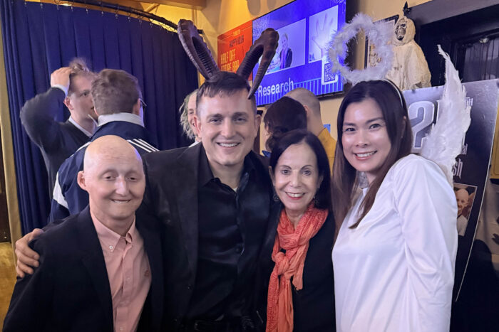 Four people have their arms around each other. One woman is wearing an angel costume and one man is wearing devil horns.