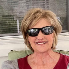 A headshot of a woman with black sunglasses. She has on a wig that is short blonde hair and she has on a red shirt.