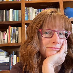 A woman wears a wig that has light red hair and she's wearing pink glasses. She's sitting in front of shelves of books.