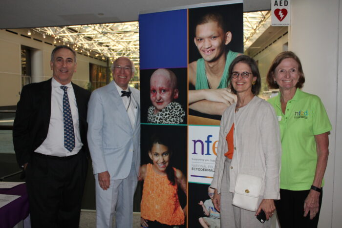 The four planners stand beside a vertical banner that says NFED and has photos of people affected by ectodermal dysplasias on it.