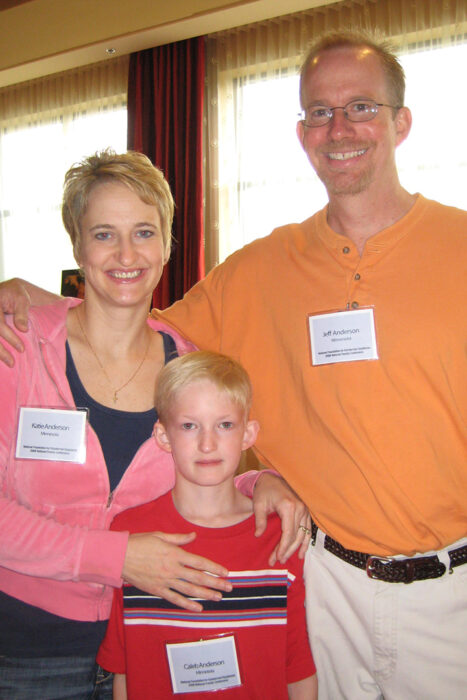 A  man stands with his arm around his wife. The wife has her arms around her son. They are standing in a hallway of a conference center.