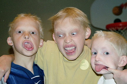 Three boys are making faces and being silly. They are sticking out their tongues. All have sparse, blonde hair. The middle boy has his arms around the boys on each side.
