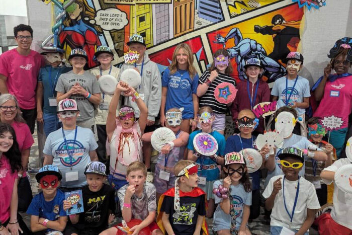 A group of kids are lined up for a photo at Kays' Kids Camp. Many are wearing baseball hats with their names on it. Some have shields like super heroes. Some are wearing masks.