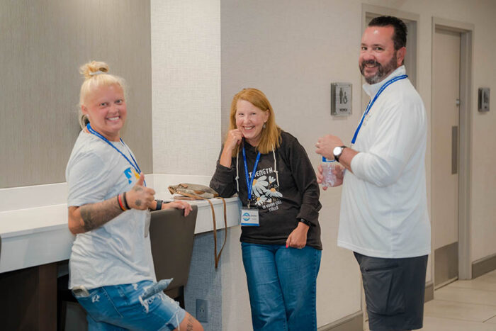 Three people are standing in a conference hallway. One woman is giving a thumbs up.