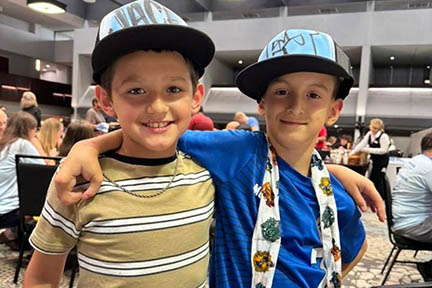 Two young boys have their arm around each other in a conference room. Each are wearing a baseball hat with their names painted on them.