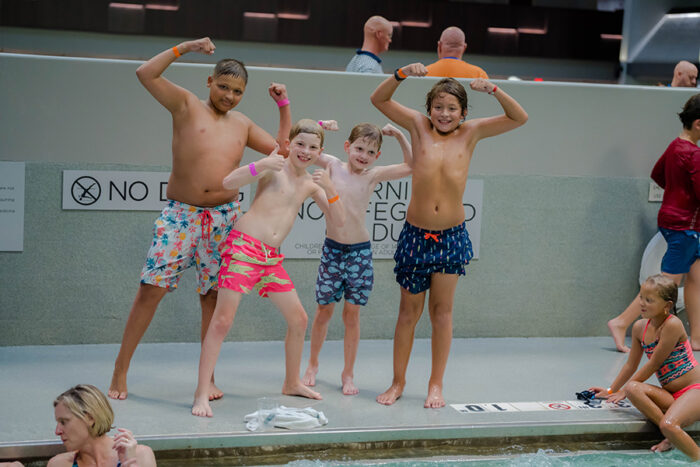 Four boys are in their swimming trunks next to a pool flexing their arms. 