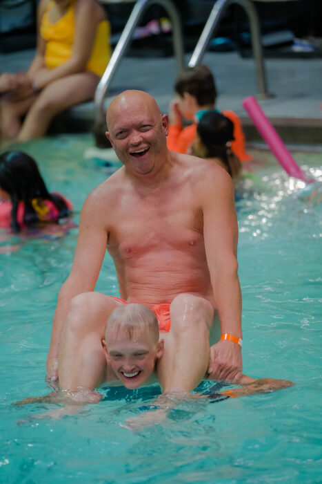 A man with XLHED is sitting on a teenagers shoulders in the swimming pool and both are laughing.