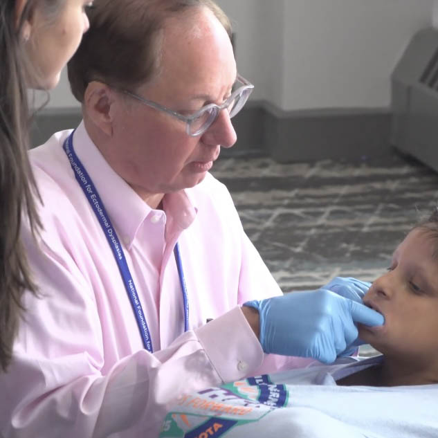 A dentist is wearing gloves and has his fingers in a child's mouth while the mother watches over his shoulder.
