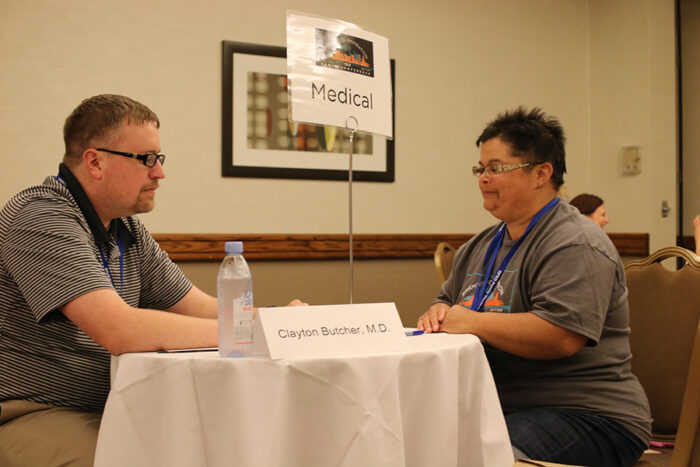 Dr. Clayton Butcher sits across a small round table from a woman. There's a sign on the table with his name and a water bottle.