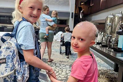 Two young girls are holding hands and walking at a Family Conference. One girl is wearing a backpack. The other girl has sparse hair and has hearing aids.