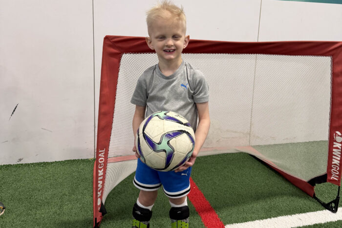 A boy with sparse blonde hair holds a soccer ball in front of a soccer goal in an indoor field. 