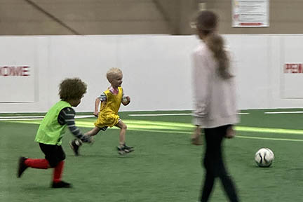 A young boy wearing a yellow uniform top runs toward a soccer ball while an opposing player in a green uniform top runs, too. A referee watches.