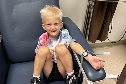 A young boy with XLHED sits in a chair in a medical room with his arm out. There's an apparatus on his left arm measuring sweat output. He's being tested for research follow-up for the XLHED therapy he received.