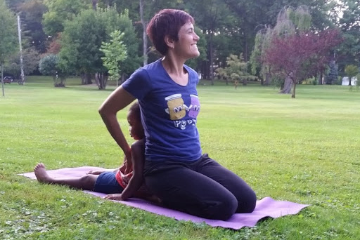 A woman is kneeling on a yoga mat in a park. A little girl is sitting back to back with her. The woman has her arm on the little girl. They are participating in a yoga class for an NFED fundraiser.