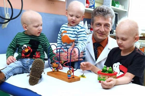 A doctor in a white coat holds one toddler as another toddler sits close by playing with toys. The older brother stands next to him also playing. All of the boys are affected by XLHED.