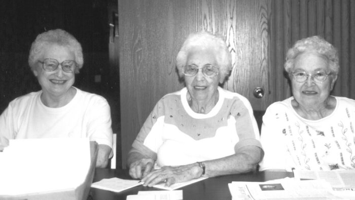 Three older women with gray hair are sitting at a table in the NFED office. They have a mailing tray and a stack of newsletters in front of them.