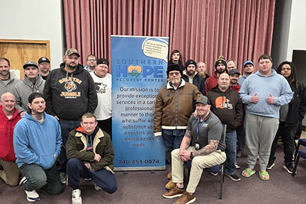 Paul sits on a chair with a large group of men behind him. There's a banner in the middle that advertises Southern Hope Recovery Center.