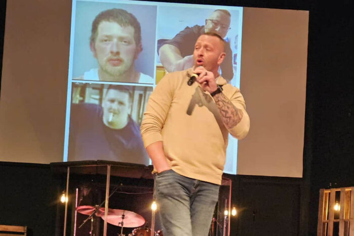 Paul stands on a stage talking into a microphone. There's a screen behind him with photos of himself on it.
