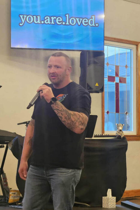 Paul is standing in front of a piano talking into a microphone. He's wearing a black tshirt and blue jeans. There's a screen overhead that says "You are loved." There's a stained glass window in the back with a cross on it.