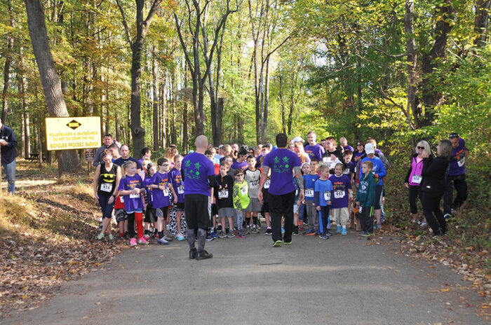 A group of children and adults are lined up on a road through a park for a fundraiser. They are all wearing racing bibs. Two men in purple shirts  are facing the front of the line getting them ready to begin.