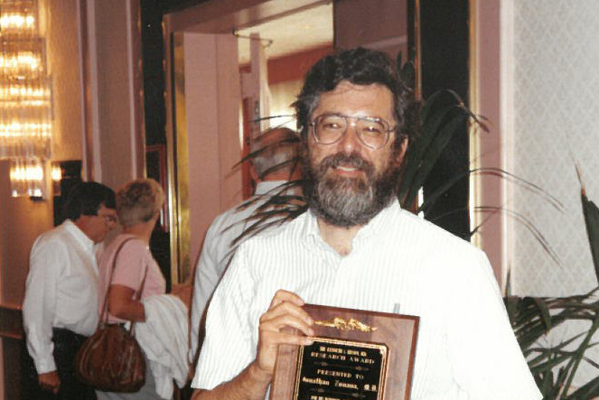A man with dark hair, glasses and a black and gray bears stands holding an NFED Research Award. People are walking behind him.