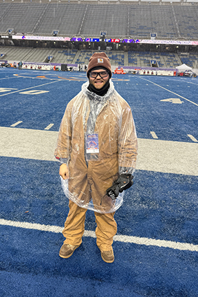 Sam is wearing rain gear and is at at large football stadium. 