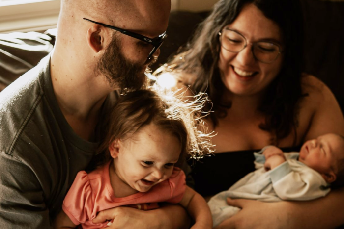 The couple sits on a couch with the dad holding the toddler daughter rand the mom holding the baby boy.