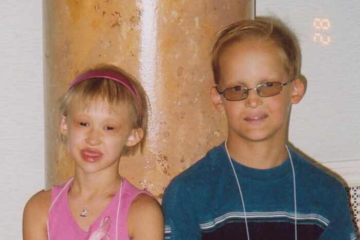 Sam is wearing glasses and stands against a column next to another young girl with XLHED. She's wearing a pink shirt and headband in her hair.
