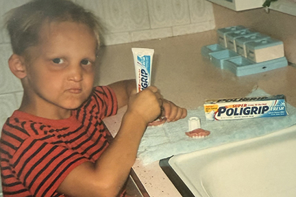 Sam as a young boy is standing in front of a bathroom sink putting Polygrip on his dentures. 