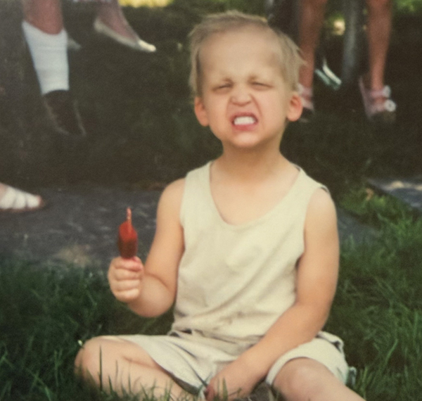 Sam as a young boy is sitting on the grass eating a red popsicle. He's smiling and you can see he's wearing his dentures. 