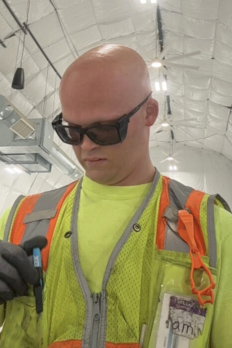 Sam is wearing  a neon yellow shirt and construction vest and black safety glasses.