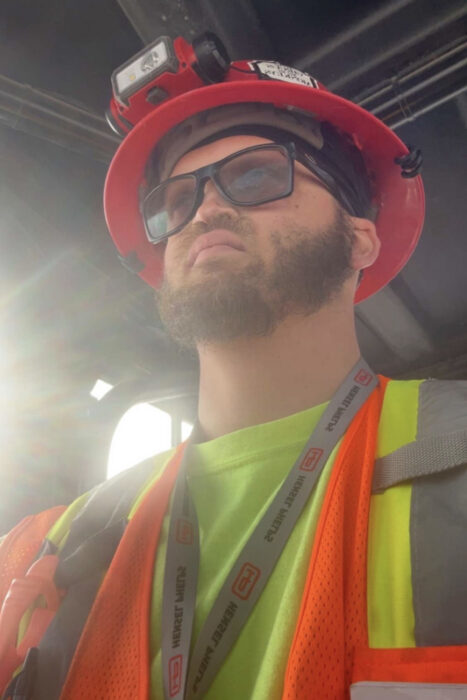 Sam is wearing a neon yellow shirt and neon orange construction vest. He has black safety glasses and a beard. He's also wearing a hard hat.