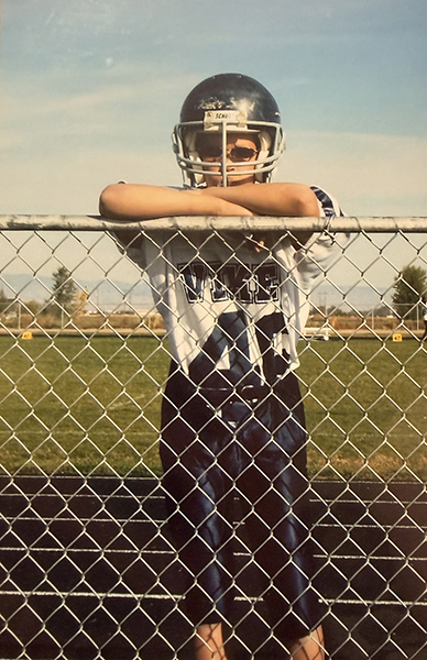 Sam as a grade schooler is in a navy and white football uniform and he's standing in a football field by a fence. 