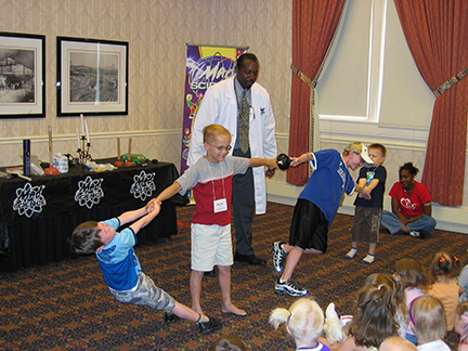 A man in a lab coat is presenting in front of a group of kids sitting on the floor. Sam stands in the middle with his arms straight out. Boys on either side of him are pulling on each arm.