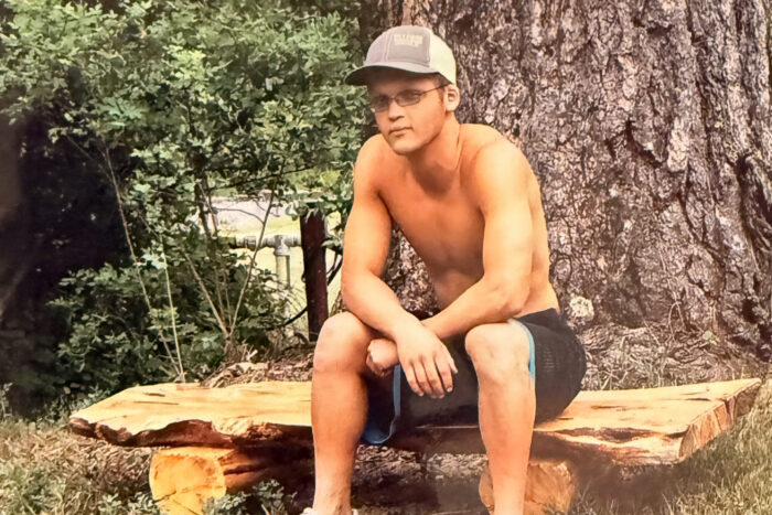 Sam as a teen sits on a log outside. He's wearing a baseball hat and glasses and has his shirt off to stay cool.