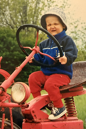 Sam is wearing a hat as a toddler and sitting on a tractor. He's wearing a blue jacket and red sweat pants. 