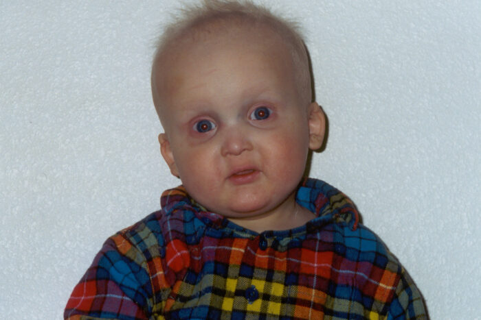 This is a head shot of Sam as a baby. He's wearing a plaid, button up shirt. He has sparse blonde hair  and big eyes.