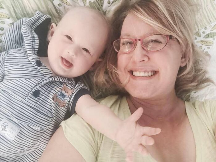 A baby affected by XLHED lays next to his mom on a bed. The boy is wearing a blue onesie. The mom has blonde hair and glasses. The boy is holding out his left arm.