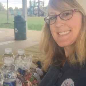 Beth is shown at a park. She's wearing an NFED t-shirt. There's a table next to her with water bottles and snacks.