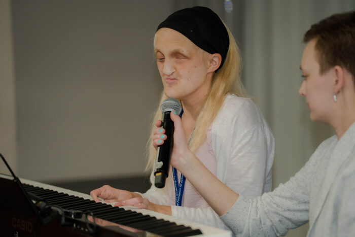 A young adult woman with blonde hair and a black headband is playing the keyboard. Lucy is holding a microphone for her to sing and speak.