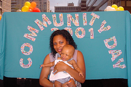 A Black mom holds her baby daughter. Behind her is a sign that says Community Day 2001. There are also balloons in the background. The baby is laughing.