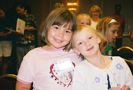 Two young girls lean on each other smiling.
