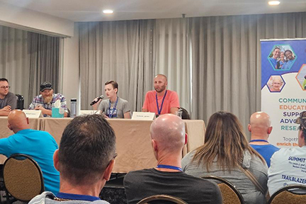Four adults are behind a table on a riser in a conference room. Lucy has the microphone and is speaking. You see the back of people's heads who are listening to her. There's an NFED banner on the right side.